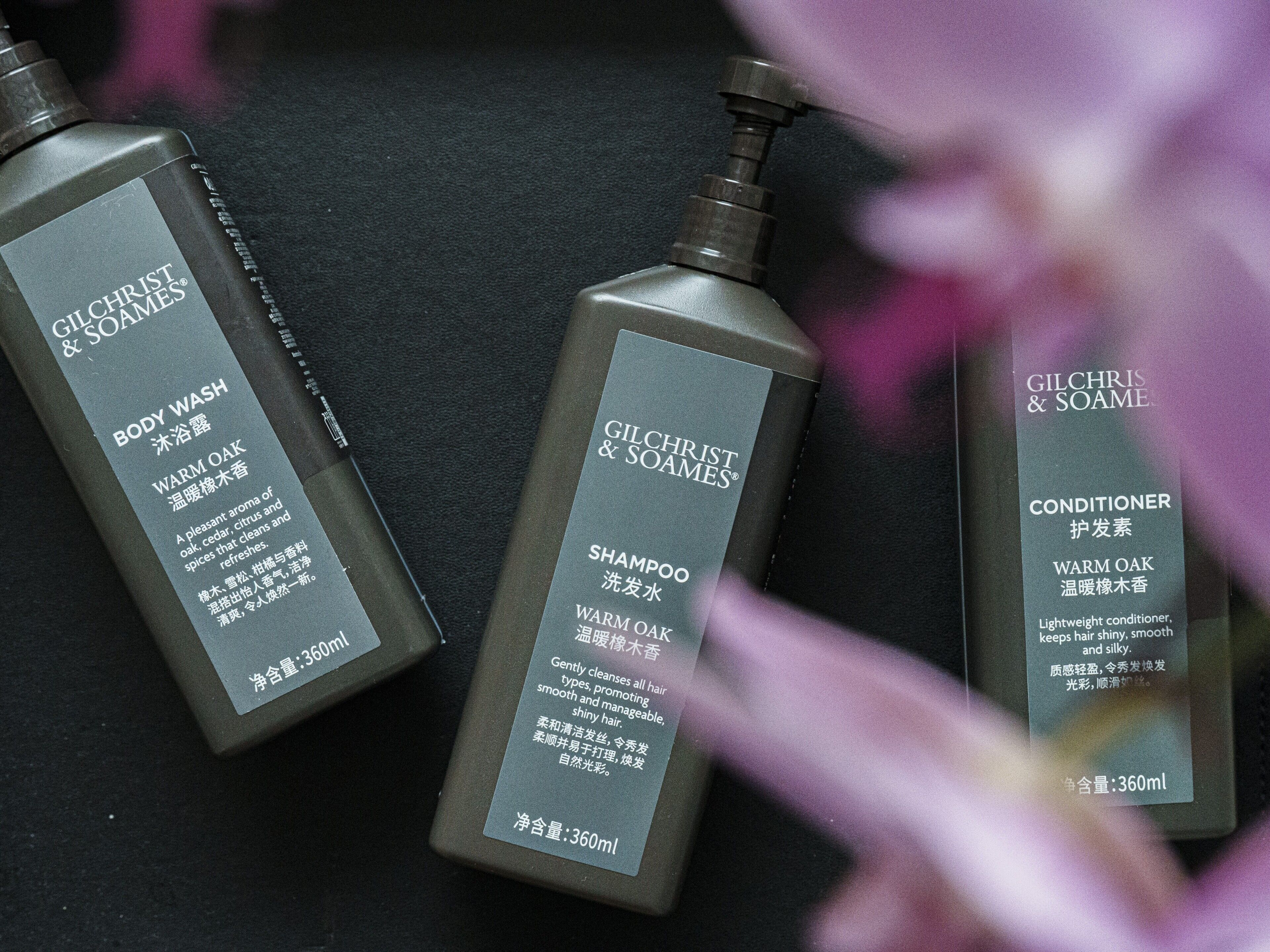 Tres botellas de crema y acondicionador para el cuerpo Gilchrist & Soames sobre una superficie oscura.