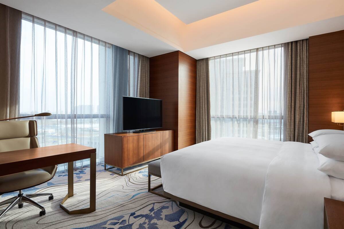 A hotel room with a large bed, a desk with a chair, a television, and a large window with curtains.