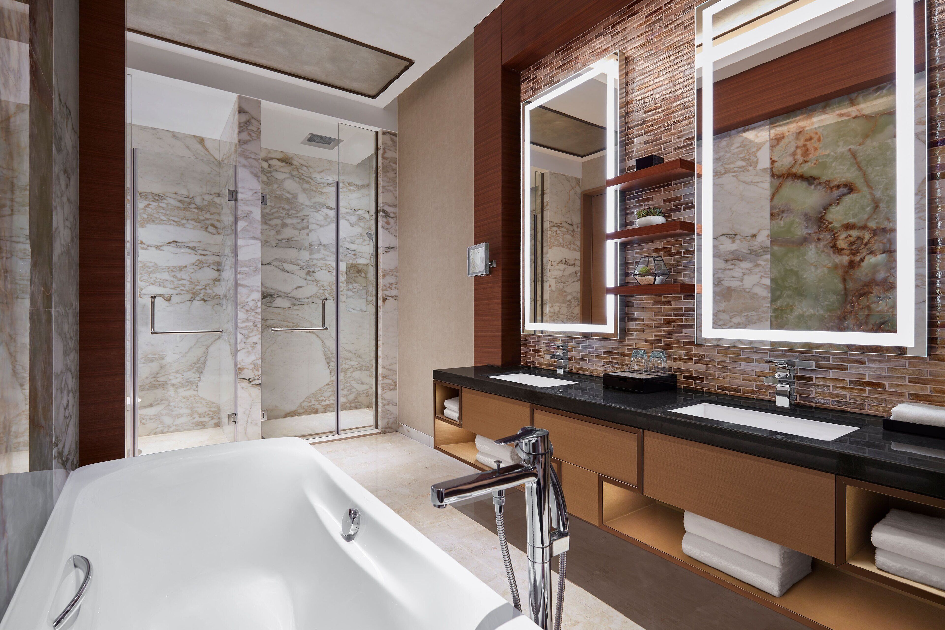 A modern bathroom with a large bathtub, two sinks, a glass shower enclosure, and a mirror with a brick backsplash.