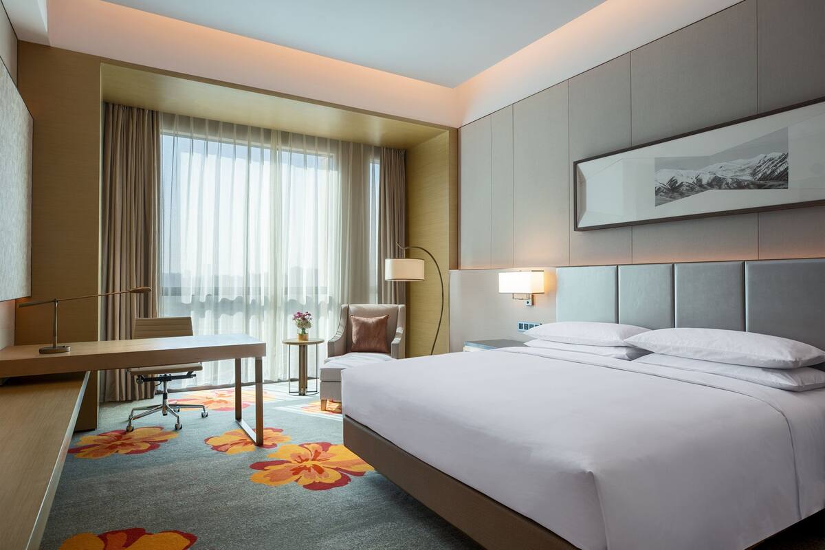 A hotel room with a bed, a desk, a chair, a lamp, and a window with curtains.