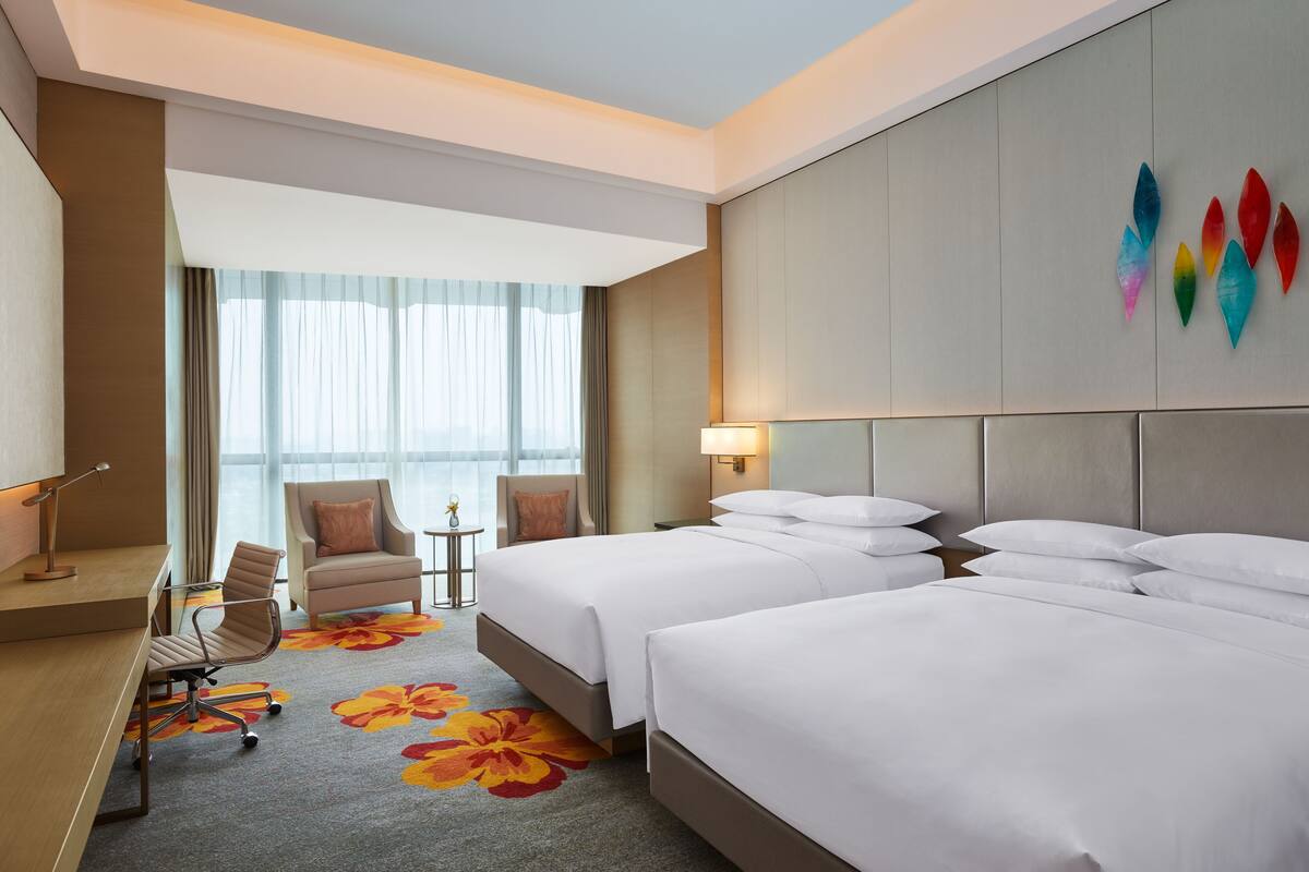 A hotel room with two beds, a desk, a chair, and a view of the outdoors.