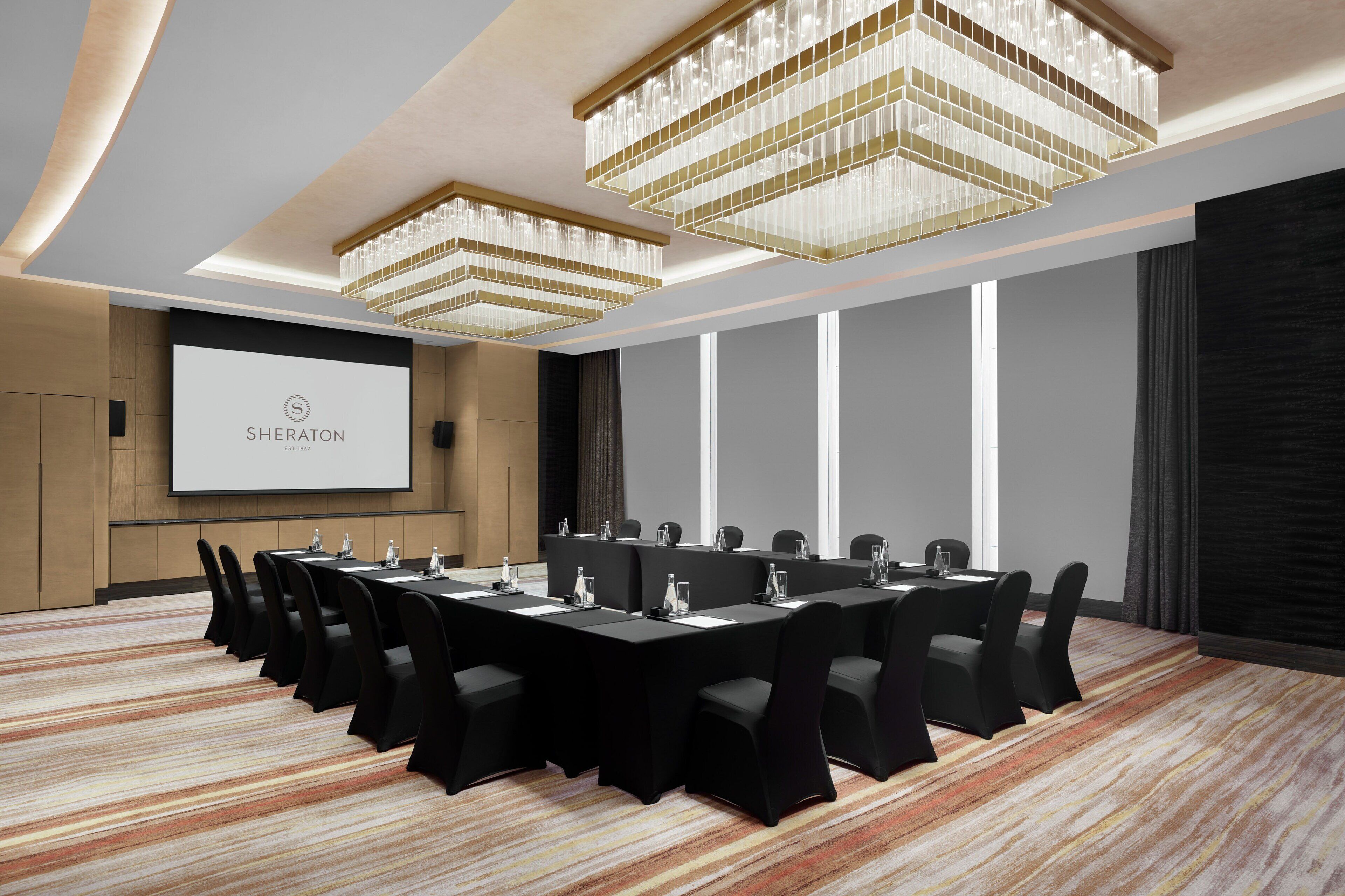 Un salón de conferencias con una larga mesa, sillas negras y una pantalla de proyector que muestra "SHERATON".
