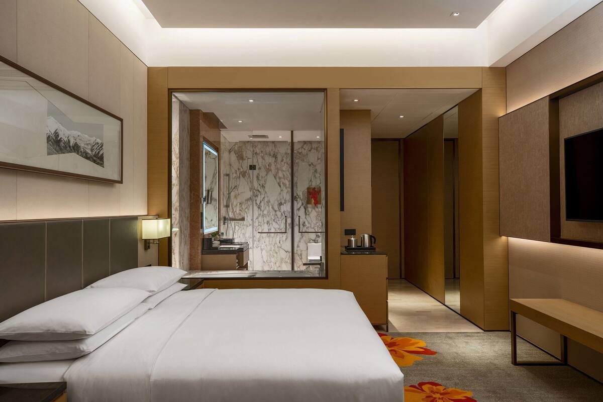 A modern hotel room with a large bed, a bedside table, a mirror, a TV, and a bathroom visible through an open door.