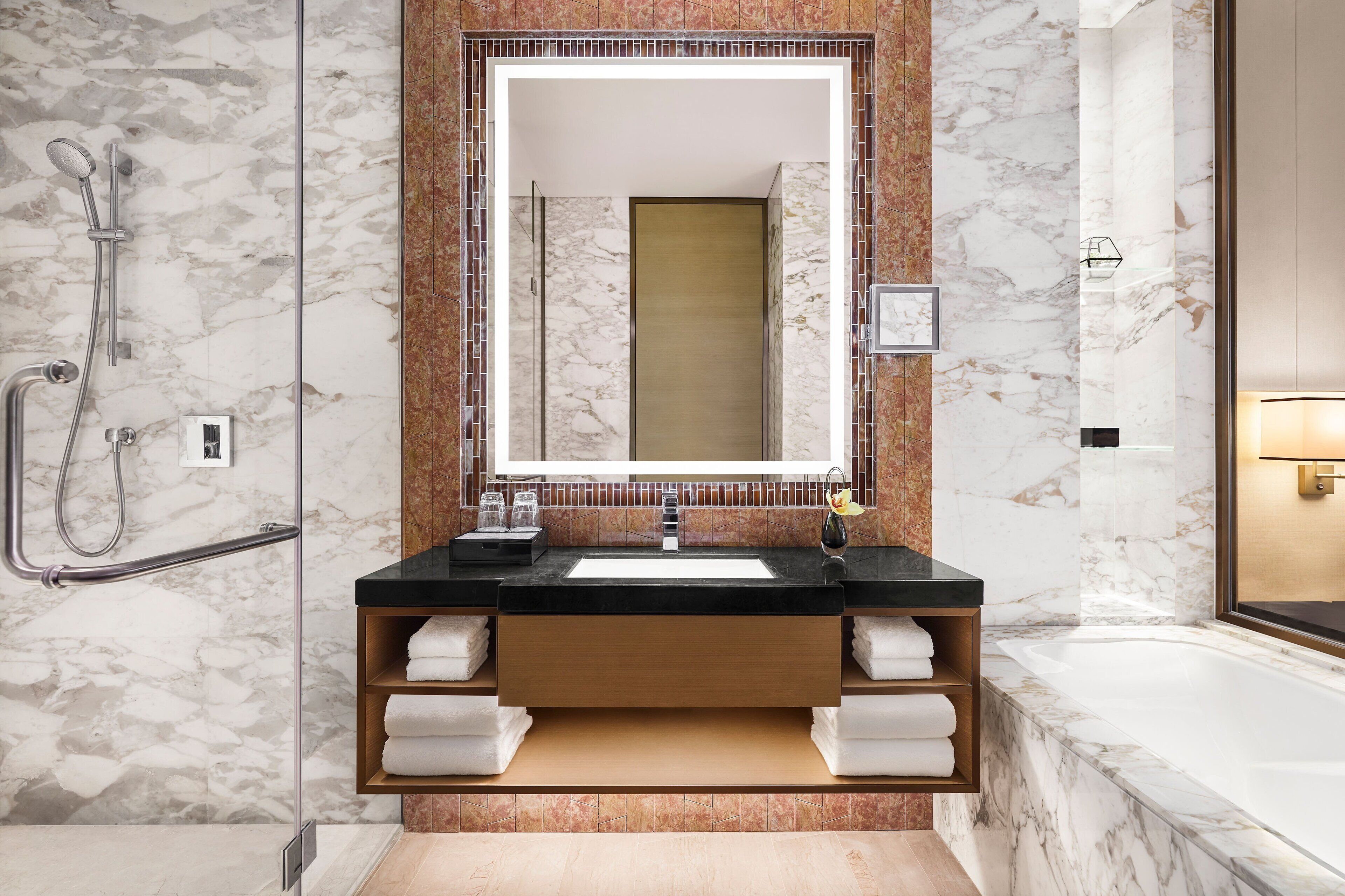 A modern bathroom with a marble wall, a large mirror, a black countertop, a wooden vanity, and a bathtub.