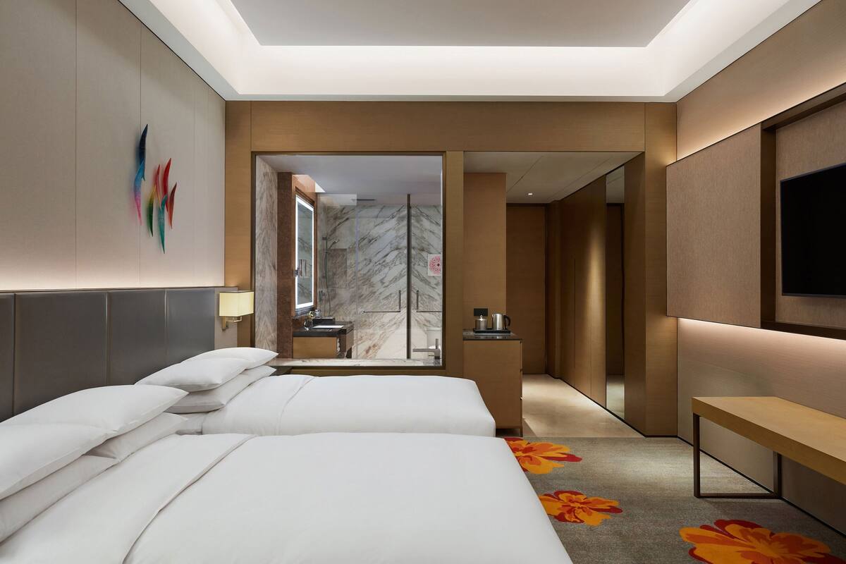 A modern hotel room with a large bed, a desk, a TV, and a bathroom visible through a glass door.