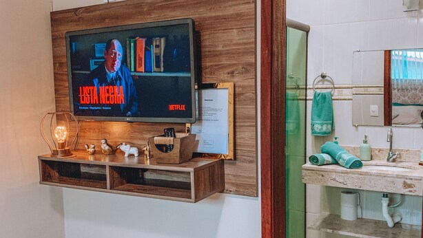 Ein modernes Wohnzimmer mit einem Flachbildfernseher an einer Holzwand, einem kleinen Hocker und einer Glastür zum Badezimmer.