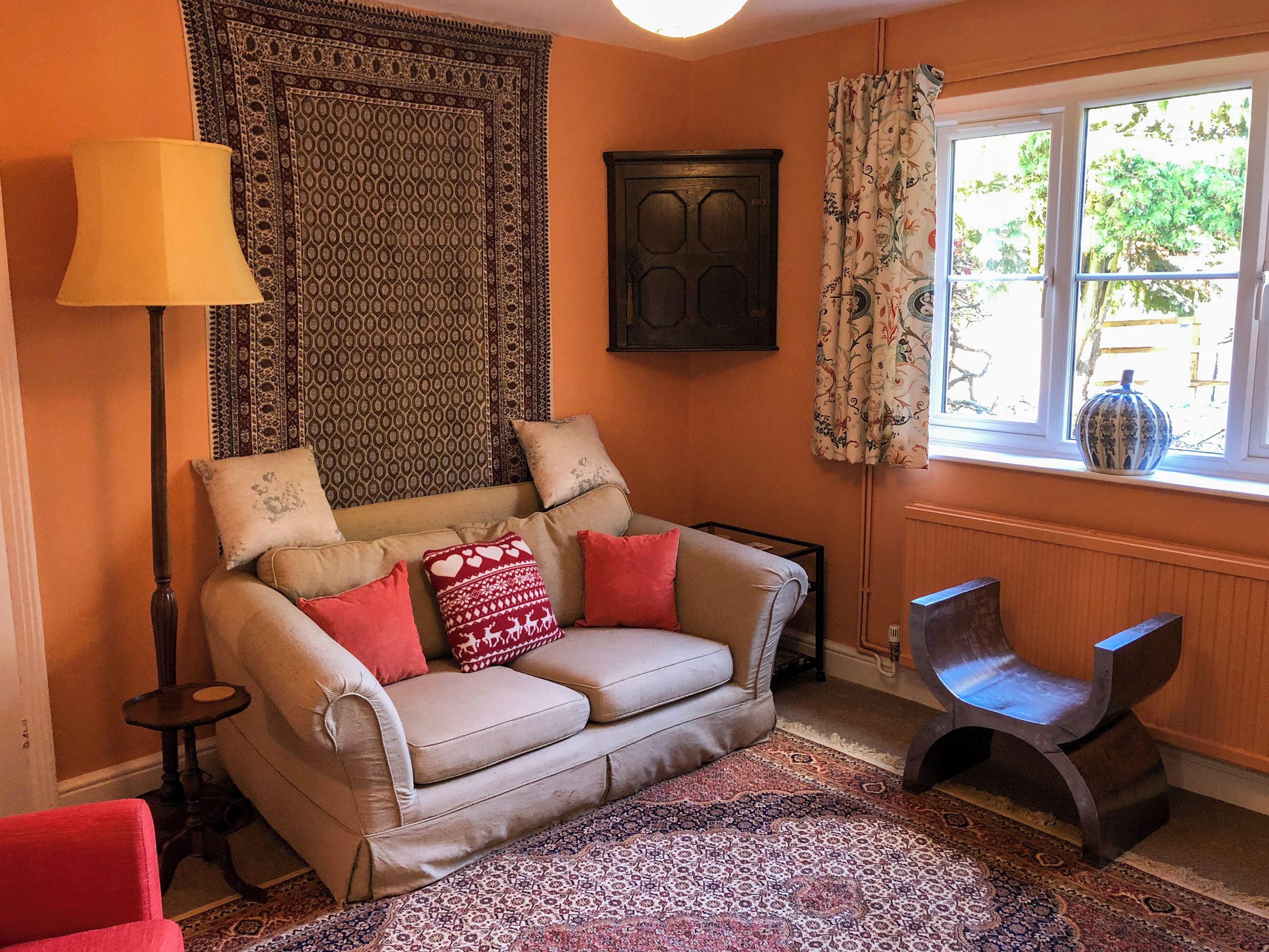 A cozy living room with a beige sofa, patterned cushions, a red armchair, a floor lamp, a patterned rug, and a window with curtains.