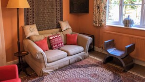 A cozy living room with a beige sofa, patterned cushions, a red armchair, a floor lamp, a patterned rug, and a window with curtains. - 4 bedroom accommodation in Hempstead, near Holt (Hempstead, near Holt)