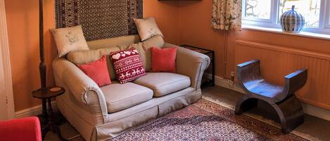 A cozy living room with a beige sofa, patterned cushions, a red armchair, a floor lamp, a patterned rug, and a window with curtains.