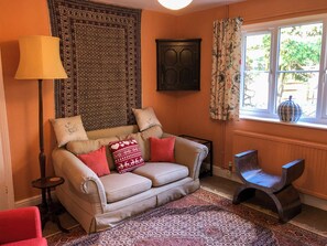 A cozy living room with a beige sofa, patterned cushions, a red armchair, a floor lamp, a patterned rug, and a window with curtains. - 4 bedroom accommodation in Hempstead, near Holt (Hempstead, near Holt)