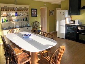 A kitchen with a dining table, chairs, a refrigerator, an oven, and a shelf with dishes. - 4 bedroom accommodation in Hempstead, near Holt (Hempstead, near Holt)