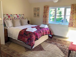 A bedroom with a bed, pillows, a nightstand, a window with curtains, and a radiator. - 4 bedroom accommodation in Hempstead, near Holt (Hempstead, near Holt)