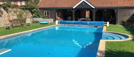 A rectangular swimming pool with a tiled deck, surrounded by a well-maintained lawn and a brick building with a tiled roof.