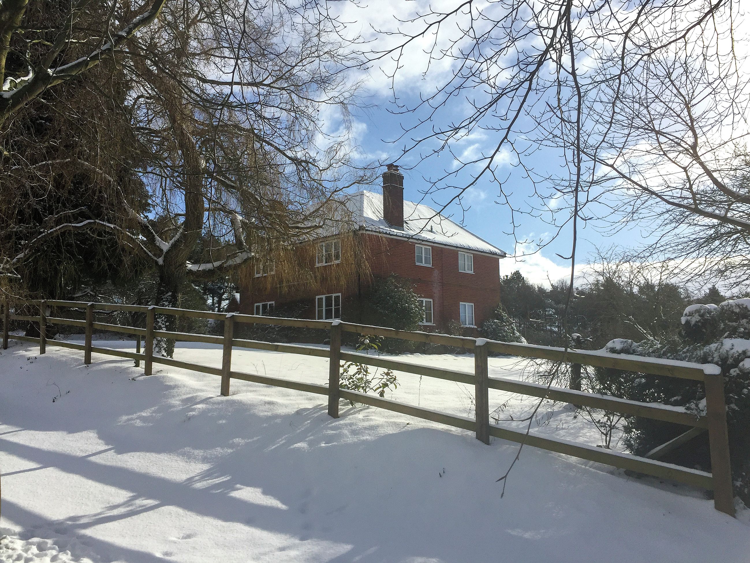 A snow-covered landscape with a brick house, a wooden fence, and bare trees.