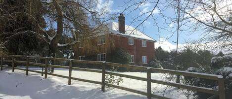 A snow-covered landscape with a brick house, a wooden fence, and bare trees.