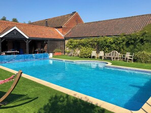 A swimming pool with a wooden deck, a covered outdoor seating area, and a brick building in the background. - 4 bedroom accommodation in Hempstead, near Holt (Hempstead, near Holt)