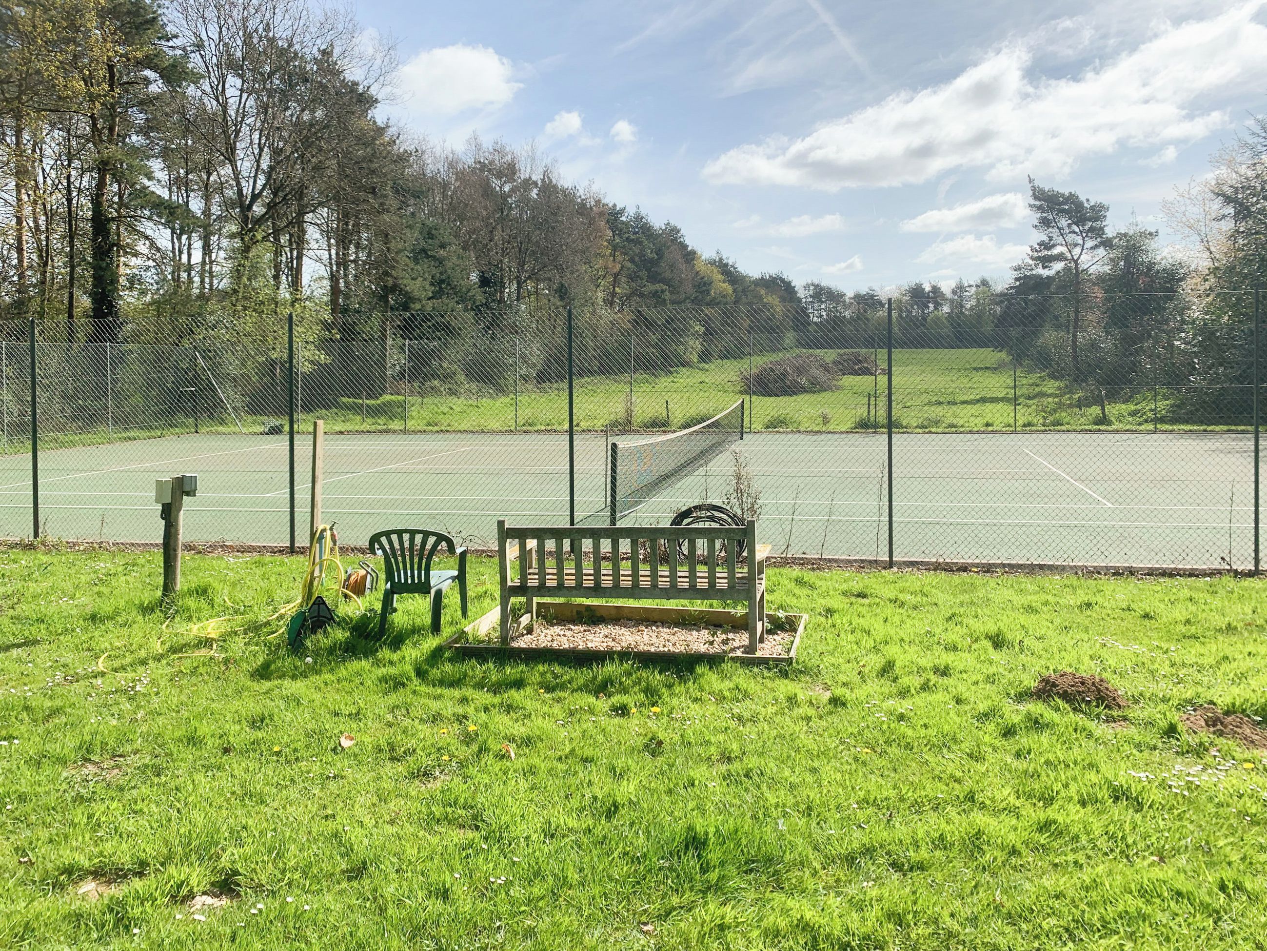 A tennis court with a bench and chairs in the foreground, surrounded by a fence and trees.