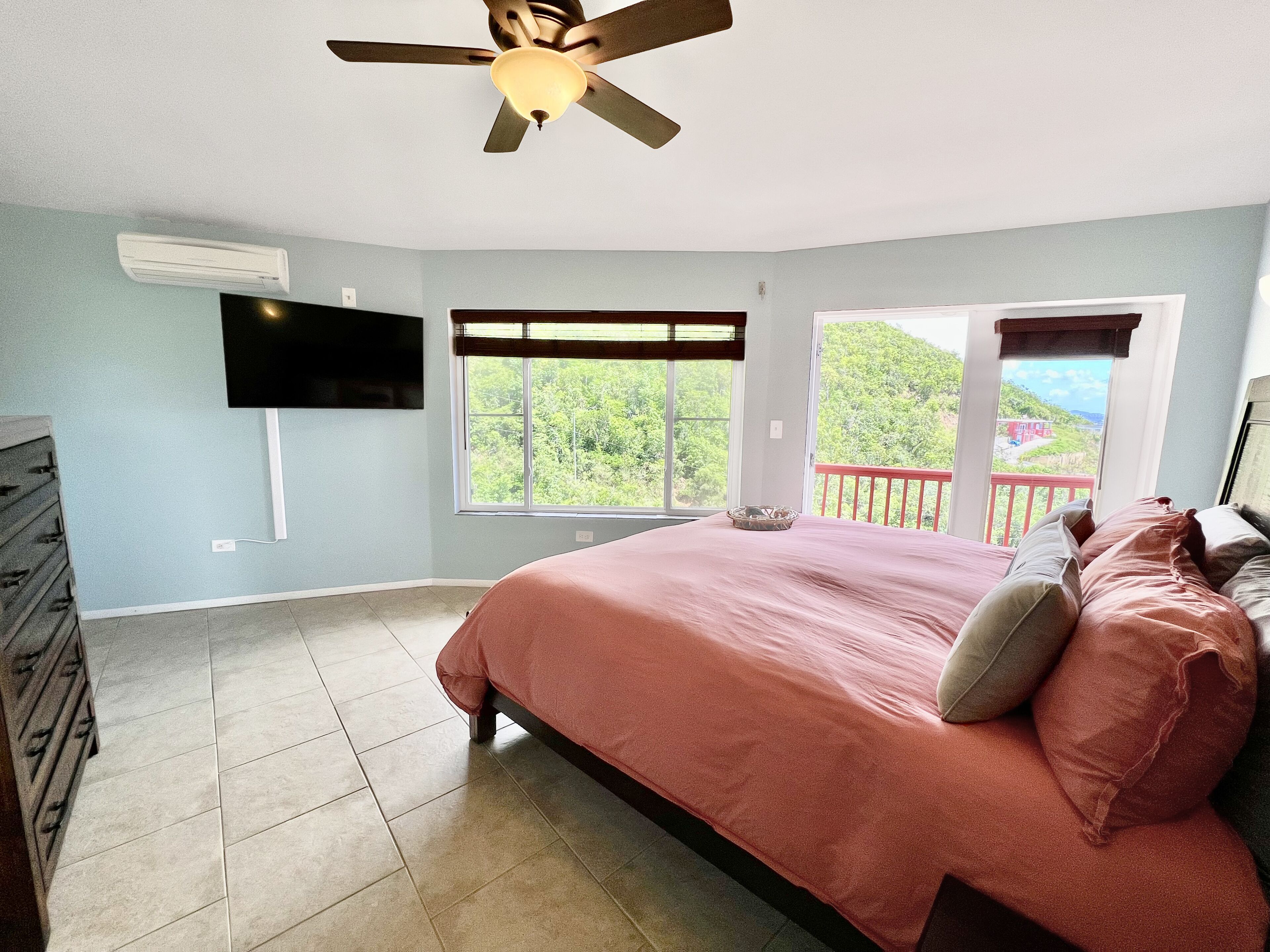 A bedroom with a bed, a ceiling fan, a TV mounted on the wall, and a window with a view of greenery.