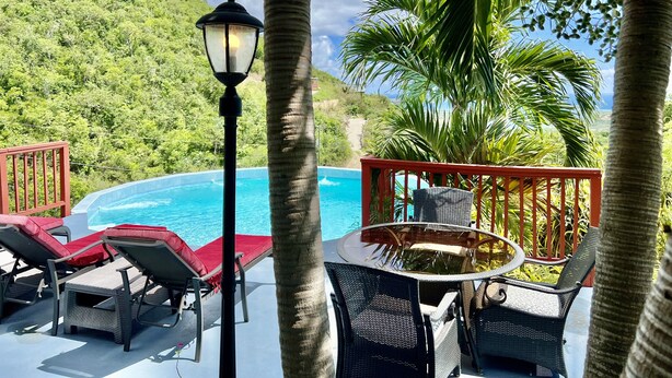 A poolside area with lounge chairs and a table, surrounded by palm trees and a view of a lush hillside. - Breathtaking Panoramic Views Of The Caribbean Sea From This Luxury Villa (St. Thomas)