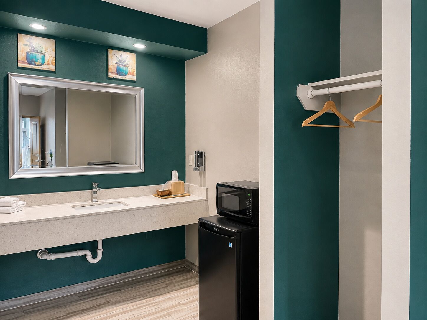 A modern bathroom with a teal accent wall, a large mirror, a sink with a towel rack, a black microwave, and a wooden towel holder.