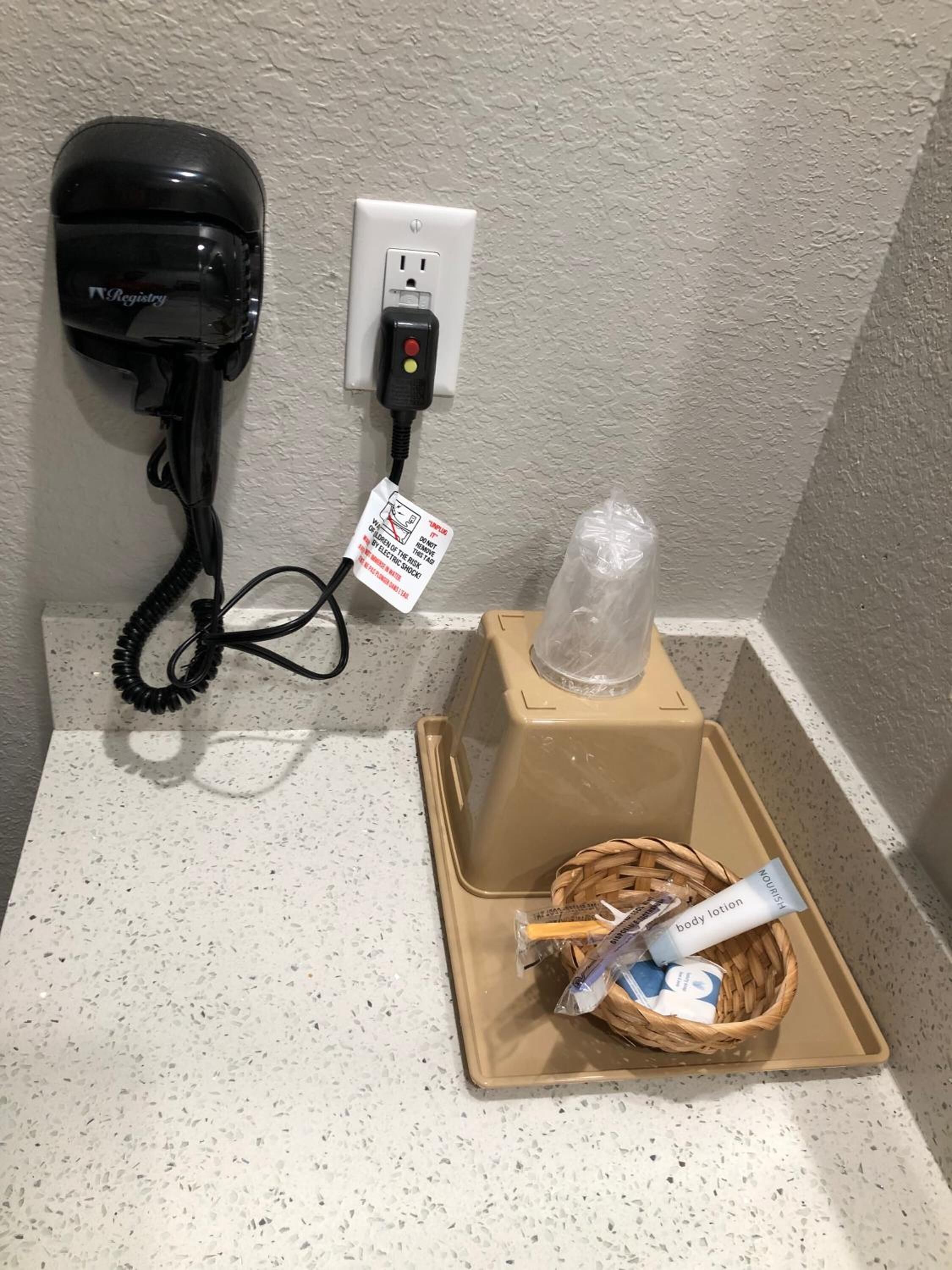 Een haardroger aangesloten op een stopcontact, een plastic fles en een mandje met toiletartikelen op een aanrecht.
