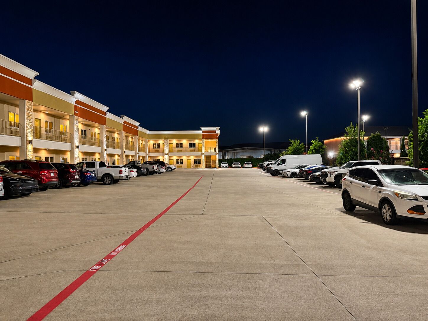 Un hotel de varios pisos con estacionamiento, mostrando una fila de coches aparcados y una línea roja que indica la salida.