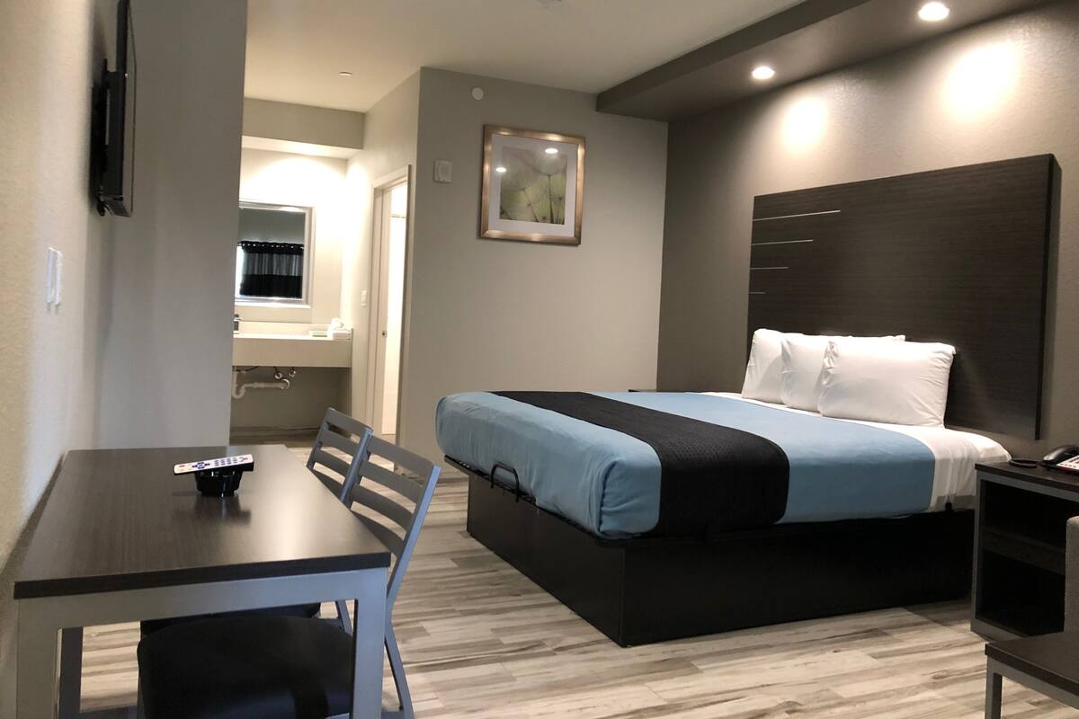 Quarto de hotel com cama, escrivaninha, cadeira, mesa de cabeceira e televisão fixada na parede.