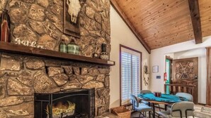 A stone fireplace with a mantel shelf, a dining table with chairs, and a wooden ceiling. - Timbers Lodge | Your Ruidoso Retreat w/ Hot Tub Bliss, Wildlife Views & Cozy Comforts (Ruidoso)