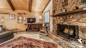 A cozy cabin interior with a stone fireplace, wooden ceiling beams, and a television. - Timbers Lodge | Your Ruidoso Retreat w/ Hot Tub Bliss, Wildlife Views & Cozy Comforts (Ruidoso)