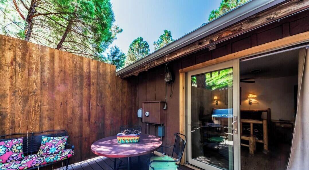 A wooden deck with a round table and two chairs, a small cabinet, and a sliding glass door leading to an interior space.