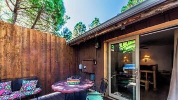 A wooden deck with a round table and two chairs, a small cabinet, and a sliding glass door leading to an interior space.
