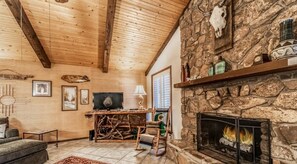 A cozy cabin interior with a stone fireplace, wooden ceiling beams, and a television. - Timbers Lodge | Your Ruidoso Retreat w/ Hot Tub Bliss, Wildlife Views & Cozy Comforts (Ruidoso)