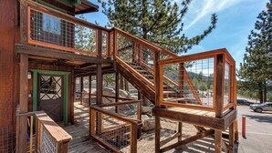 Una scala in legno conduce a un terrazzo con struttura in vetro, immerso tra alberi e sotto un cielo sereno.