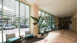 Relax in the spacious lobby with large windows offering a view of the street, comfortable seating areas, and a lush potted plant.