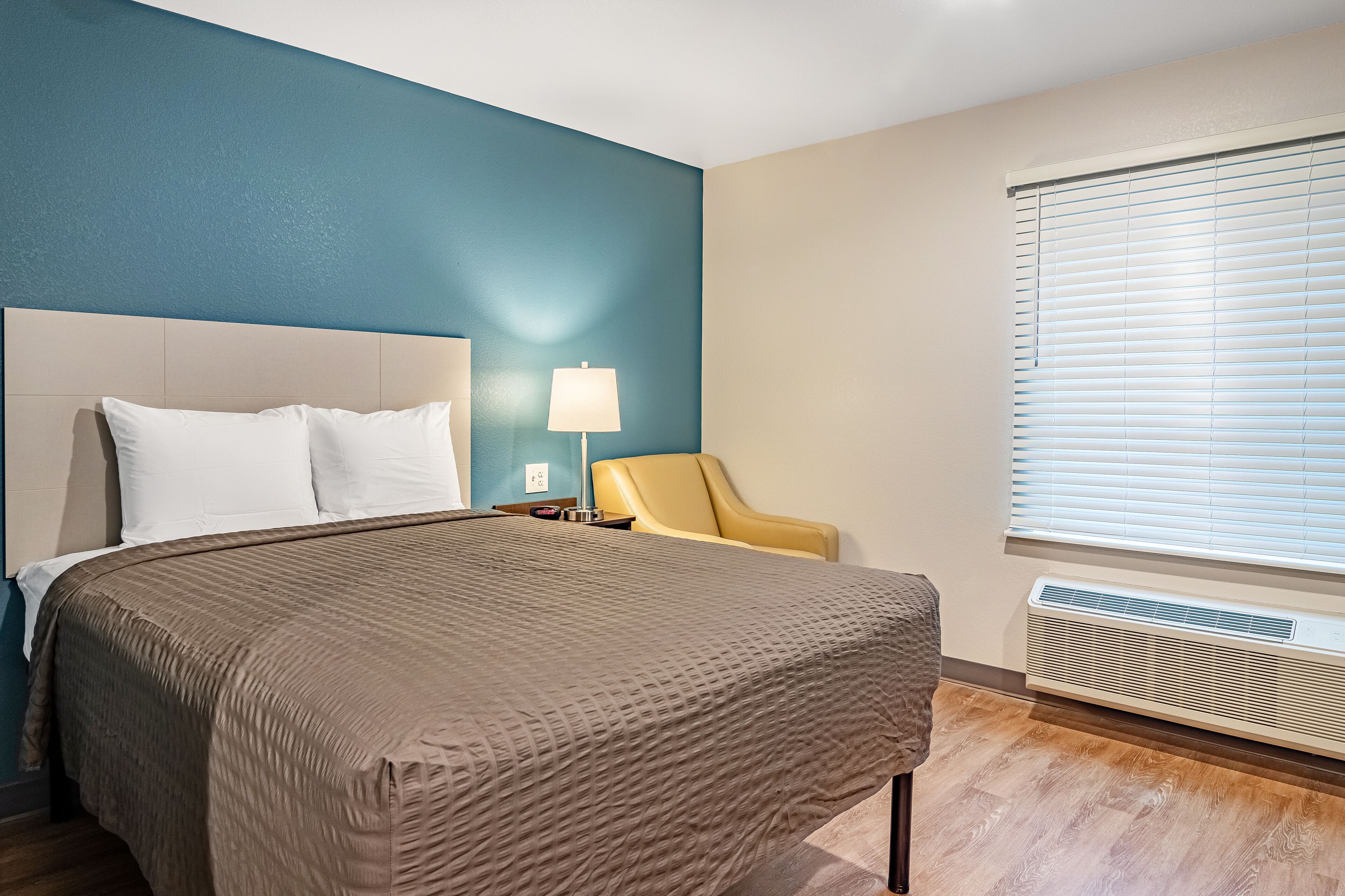 A hotel room with a bed, a chair, a lamp, and a window with blinds.