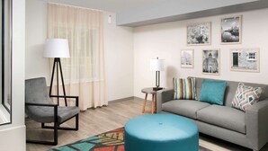 A modern living room with a grey sofa, a blue ottoman, a black chair, a floor lamp, and a patterned rug. - WoodSpring Suites Toledo Maumee (Maumee)