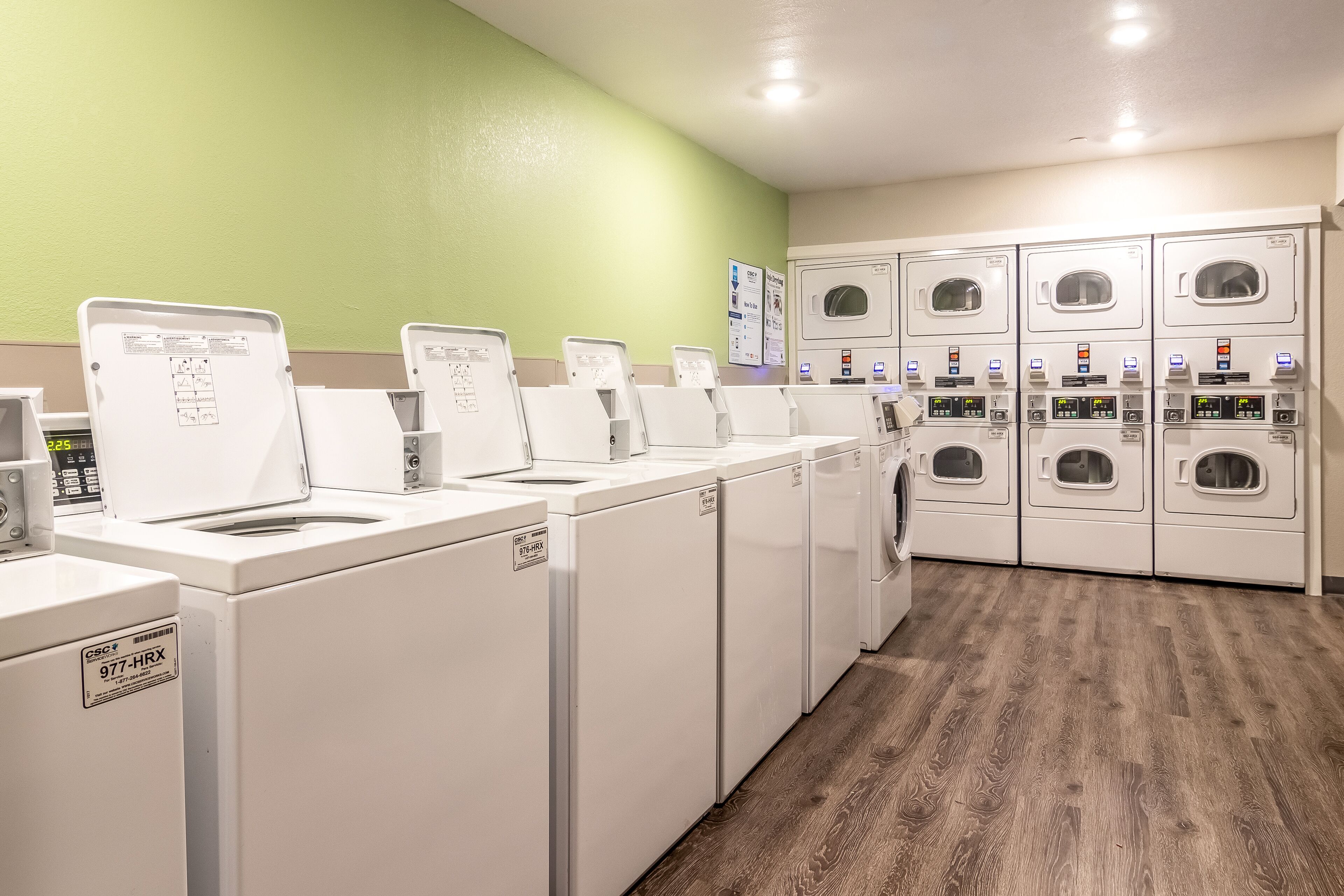 A laundry facility with multiple washing machines and dryers, a green wall, and a wooden floor.