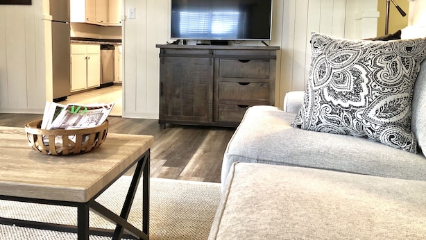 Una sala de estar con un sofá, una mesa de centro con revistas, un televisor en un mueble y una cocina al fondo.