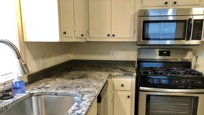 Una cocina con pileta de acero inoxidable, encimera de granito, alacenas blancas, microondas integrado, estufa y horno.