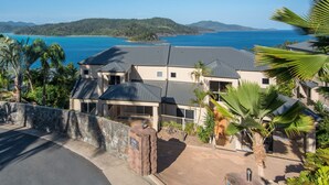 A multi-story house with a stone wall entrance, a driveway, and a view of the sea and hills in the background.