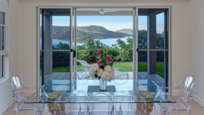 A modern dining area with a glass table and chairs, a vase of flowers, and a scenic view of a lake and hills through large glass doors.