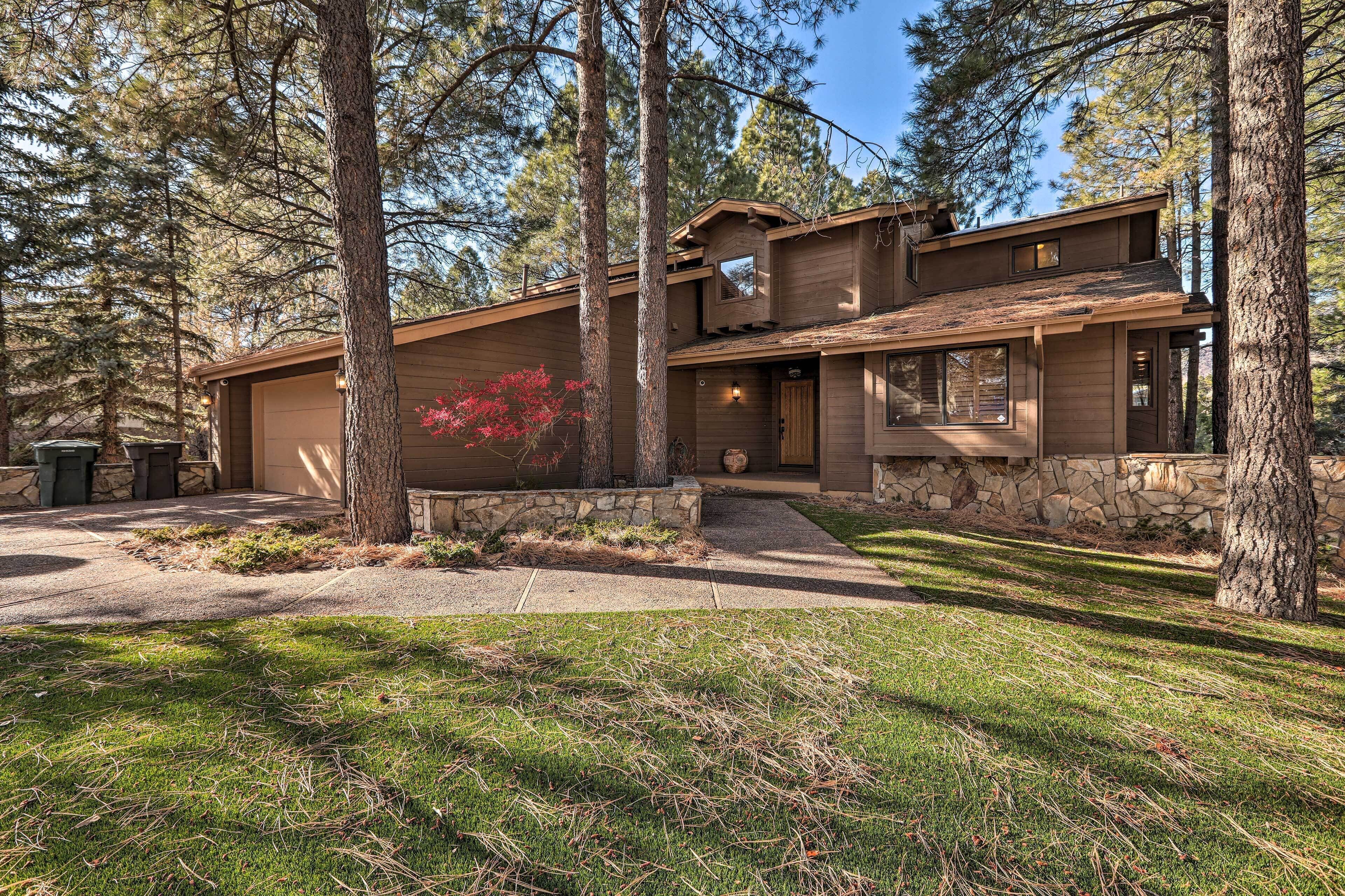 A two-story wooden house with a stone foundation, surrounded by tall trees and a well-maintained lawn.