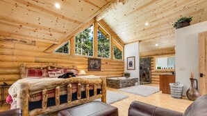 L’intérieur d’un chalet en rondins, avec un lit, des sièges en cuir, une cheminée en pierre et une vue sur l’extérieur.
