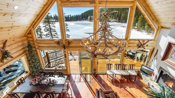 Une cabane en rondins dotée d’une grande fenêtre en verre qui offre une vue sur un paysage enneigé et une rivière. À l’intérieur, on trouve une table à manger en bois avec des chaises, une cheminée et un téléviseur.