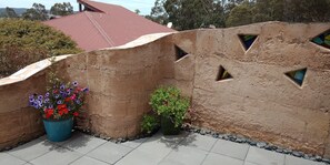 A terracotta wall with geometric cutouts, a potted plant with red and purple flowers, and a red-roofed building in the background. - Gingko Lodge. Country luxury with a view. (Mossiface)