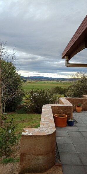 A patio area with a stone bench, potted plants, and a view of a green landscape under a cloudy sky. - Gingko Lodge. Country luxury with a view. (Mossiface)