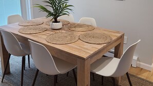 Een eethoek met een houten tafel, witte stoelen, geweven placemats en een potplant.