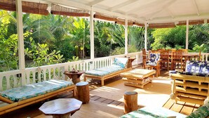 A covered outdoor seating area with wooden furniture and a view of lush greenery. - Villa Iris by Tahiti Homes  (Moorea-Maiao)