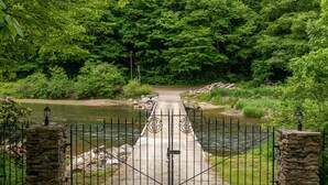 Un sentier bordé de grilles mène à une rivière, entourée d’une végétation luxuriante et de piliers en pierre surmontés de lanternes.