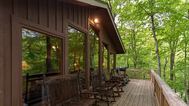 Une terrasse en bois, dotée de fauteuils à bascule et d’un barbecue, entourée d’arbres.